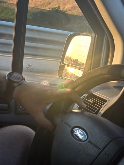 The image, taken inside a vehicle, shows a person's hand on the steering wheel, which has a Ford logo. A smartwatch on the person's wrist displays the time "12:56". The sun is setting, casting a warm glow and creating lens flares, visible in the side mirror which reflects the sky and distant trees. The vehicle's interior, including an air vent, is also visible. The environment outside the vehicle appears to be a road with a guardrail and vegetation. There is no specific indication of the location being the city of Szeged, Hungary.