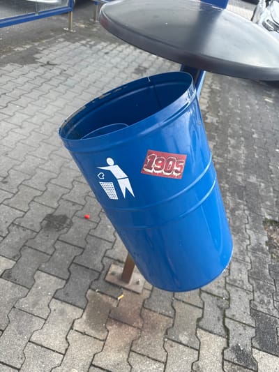 A blue metal trash can with a black lid stands tilted on a pole on a gray paved walkway. A white silhouette of a person throwing an object into a trash can is visible on the side of the trash can, along with a red sticker that reads "1905". The ground is wet in some areas, and there is a cigarette butt on the ground near the base of the trash can pole. In the background, a portion of a building with blue framing and a car are visible. The scene is outdoors in an urban environment, likely a public space.