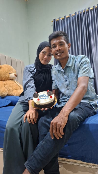 In a bedroom in Pie, Indonesia, a couple sits on a bed with a large teddy bear to their left. The woman, wearing a black hijab and a blue blouse, holds a round cake with white frosting decorated with cookies and cherries. The man, in a patterned blue shirt and jeans, sits beside her, his arm around her. Both are smiling and looking towards the camera. The bed has a blue bedspread, and dark blue curtains hang in the background.