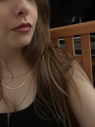 A close-up, low-angle shot shows a woman's face and part of her body. Only her nose and lips are in full focus. She has fair skin and light brown hair, with some strands covering her face and chest. A thin gold necklace is visible on her chest. She is wearing a black top. The background includes a wooden chair and a blurred dark object. No location information about Truchtersheim, France can be determined from the provided image.