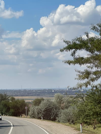 A wide shot captures a rural landscape under a bright, cloudy sky. In the foreground, a paved road curves to the left, with white lane markings. On the right side of the road, there are small white posts with red tops and sparse vegetation. A traffic sign, triangular with a red border and a black arrow pointing right, is visible. Lush green trees dominate the mid-ground, with some silvery-green foliage interspersed. Beyond the trees, a hazy horizon reveals a distant urban or industrial area with several tall, slender structures resembling smokestacks or communication towers. The sky is filled with large, fluffy white clouds against a pale blue backdrop. A portion of a white vehicle is visible on the left side of the road in the lower left corner. The scene appears to be located in or near the city of Praskoveya, Russia, given the implied context.