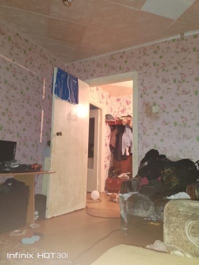 The image depicts an interior scene within an apartment in Lisakovsk, Kazakhstan. The room is cluttered and appears lived-in, with furniture and personal belongings strewn about. The wallpaper features a repeating pattern of small floral motifs and what appear to be stylized animals or figures.
In the foreground, a couch or armchair is visible, covered with various items of clothing and fabric, some of which are plastic-wrapped. To the left, a table or desk holds a dark-colored monitor and more scattered items.
A doorway leads to another part of the apartment, revealing a narrow hallway or room with more wallpaper. Inside this area, a piece of furniture, possibly a shelf unit or dresser, is laden with folded clothes and jackets hanging on the side.
The flooring appears to be a light-colored wood or laminate. A white, round object, possibly a balloon or a small stool, is positioned on the floor near the doorway.
The overall lighting is dim and appears to be artificial, casting shadows and contributing to the somewhat disorganized appearance of the space. A watermark at the bottom of the image indicates it was taken with an Infinix HOT 30i phone. No individuals are visibly present in the scene.