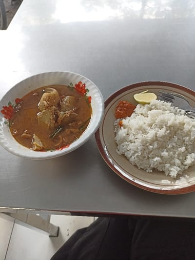 A white table with two plates on it, one with white rice, a red chili sauce and a wedge of lime, the other with a brown stew. The background is blurry and shows a person's black clothing and a tiled floor. The coordinates provided correspond to an area within the Kecamatan Waru district of Sidoarjo, Indonesia. This suggests the food is likely Indonesian and being eaten at a local restaurant.