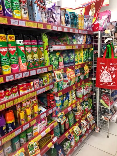 The image shows a retail store aisle in Putat, Indonesia, stocked with various household cleaning and pest control products. The shelves are filled with brands like Baygon, HIT, Kismo, Sunlight, SOS, Dahhila, and VAPE. Products include insecticide sprays, laundry detergents, dish soaps, and air fresheners. Prices are displayed in Indonesian Rupiah (Rp). A red reusable shopping bag with a cartoon drawing of a sad garbage bag and the text "Alfagift" is hanging on the right side of the image. The store appears to be well-lit and organized. There are no people visible in the image.
