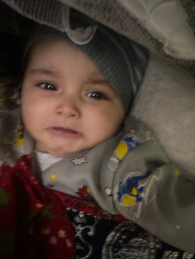 An infant is depicted in a close-up, top-down view, centered in the frame. The infant wears a grey ribbed knit hat and a grey long-sleeved garment adorned with yellow, blue, red, and white cartoon-like patterns. The child's face exhibits a distressed expression, characterized by slightly downturned lips and open eyes looking towards the camera. A light-colored, textured fabric partially obscures the top of the frame. To the infant's left and below, a red patterned fabric is visible, and further down, a dark fabric with silver embroidery is present. The environment appears to be an indoor setting. This scene is located in the city of Galaosiyo, Uzbekistan.