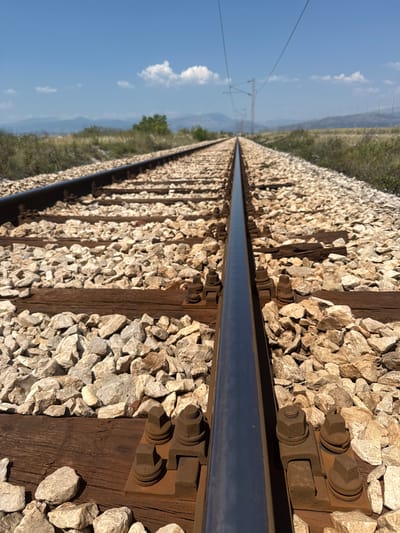 The image shows a close-up, low-angle view of a railway track extending into the distance. The tracks are made of steel rails laid on wooden sleepers and covered with light-colored gravel. The perspective emphasizes the parallel lines of the tracks and the ballast. In the background, the landscape consists of dry grass, bushes, and distant hills under a clear blue sky with a few scattered white clouds. Overhead power lines for electric trains are visible, suggesting an active railway line. The scene is devoid of any people or vehicles. The location is the city of Mojanovići, Montenegro.