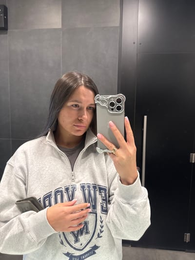 An indoor mirror selfie shows a young woman looking directly at the camera. The image is taken at a 90-degree angle, with the subject's head tilted to the right. She has dark, straight hair, parted down the middle, with a subtle blue tint visible in some strands near her face. Her expression is neutral, and her skin shows a few freckles or minor blemishes.
She is wearing a light grey, long-sleeved sweatshirt with a quarter-zip collar, revealing a darker grey or black t-shirt underneath. The sweatshirt features a prominent dark blue graphic on the front: a crest or shield design with a large "M" in the center. Above the "M," the word "MIAMI" is visible, and below it, "WE." Along the right side of the crest, oriented vertically, the words "STUDENT LIFE" are printed.
The woman is holding a smartphone, likely an iPhone, with her right hand, which is adorned with several rings on one finger. The phone has a distinctive clear or silver-toned case with wavy edges around the camera module and along the sides. Her fingernails have a classic French manicure with light pink polish and white tips. Her left hand is resting casually on her stomach, also showing the French manicure.
The background consists of dark, possibly charcoal grey, tiled or paneled walls with horizontal seams. In the upper portion of the background, a very dark, almost black, shiny surface (possibly a door or another wall section) is visible. A small, dark rectangular object is present on the lower left wall, possibly a dispenser or sensor. The lighting appears even and artificial, suggesting an indoor setting like a restroom or hallway.