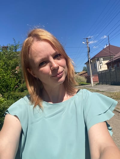 A fair-skinned woman with light red hair is taking a selfie outdoors. She is wearing a light blue-green ruffled top. The background shows a bright blue sky, green foliage, a telephone pole with wires, and houses with brown roofs. A dirt road is visible on the right side of the image. The location is the city of Zapadny, Russia.