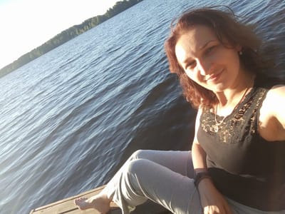 A woman is sitting on a wooden dock by a body of water. She is wearing a black top with lace detailing and light-colored pants. Her right foot is visible, resting on the dock. The water behind her is rippled, and a treeline is visible in the distance. The scene appears to be outdoors, with natural lighting. This image was taken in Suonenjoki, Finland.