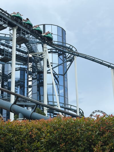 A roller coaster train with passengers is captured in motion on a track in front of a modern glass building. The roller coaster cars are green, and several individuals are visible in them, some appearing to wave. The building features a curved, reflective glass facade, with its structure visible through the transparent surfaces. The scene is set against a cloudy sky. In the foreground, a dense green hedge with some reddish-tinged leaves is present. The overall impression is that of a vibrant amusement park environment.