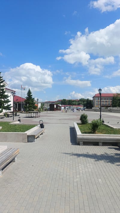 An outdoor daytime photograph captures a public plaza in Kuybyshev, Russia, on a clear day with scattered clouds. The plaza is paved with light gray rectangular bricks, and several modern wooden benches with concrete supports are strategically placed throughout. Green grassy areas with small bushes are integrated into the plaza design, bordered by concrete planters.

On the left side of the image, a white building with a red roof and dark trim is visible, featuring exterior lighting fixtures. Several flags on tall poles stand in front of this building. Evergreen trees are planted on either side of the building's entrance.

In the background, a fountain with multiple jets is active, with a few people gathered around it. Beyond the fountain, more buildings with red roofs are visible, along with trees and streetlights. A sign with Cyrillic lettering, reading "КУЙБЫШЕВ", is prominently displayed on a building in the middle ground. A lamppost with a dark black pole and a traditional black lantern is positioned in the foreground on the right, casting a shadow on the pavement. A few pigeons are visible on the ground near the lamppost.