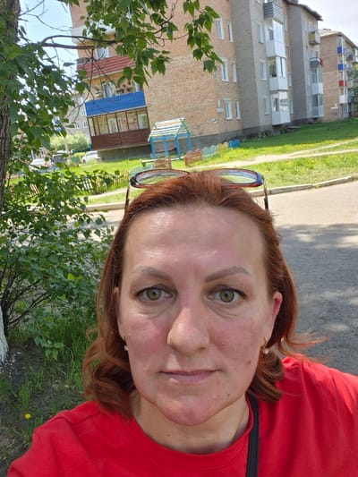 A selfie photograph captures a woman in the foreground with an urban residential environment in Kansk, Russia, in the background.
The primary subject is a woman with fair skin, green eyes, and reddish-brown hair, parted and pulled back with a pair of eyeglasses resting on top of her head. She is wearing a red short-sleeved t-shirt and small hoop earrings with clear stones. Her face appears slightly flushed or sun-kissed, with visible pores and minor skin texture. Her expression is neutral.
The background reveals a sunny day with clear skies. To the left, a tree trunk with green leaves and branches is prominent, partially obscuring the view. Beyond the woman and the tree, there is a grassy area with scattered yellow dandelions. A low, colorful picket fence (featuring blue, yellow, and red sections) separates the grassy area from an asphalt road or pathway.
Several multi-story apartment buildings, characteristic of Soviet-era architecture, dominate the midground and background. The building directly behind the woman is brick, with various types of balconies; one is enclosed with a red roof, and another below it has a blue roof and enclosed windows. Further to the right, another brick building is visible, featuring multiple grey and white enclosed balconies. In the distance, additional buildings with similar architectural styles and colored balconies (red and blue) can be seen. A small, blue and green structure, possibly part of a children's play area, is visible in the grassy area between the fence and the buildings. White cars are parked or driving in the distance on what appears to be a street.