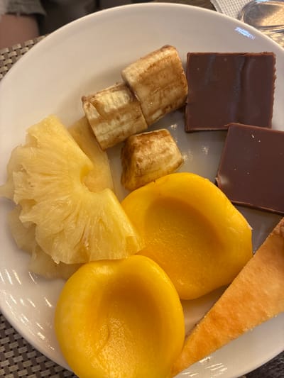 A white plate holds a selection of food items. In the upper left corner, two square pieces of dark chocolate are visible. To their right, a triangular slice of a golden-brown pastry rests. The center and right side of the plate are dominated by several pieces of canned yellow peaches, their smooth, rounded surfaces gleaming. In the lower half of the plate, slices of pineapple are arranged, their textured surfaces clearly showing the fruit's fibrous structure. Between the chocolate and the fruit, several pieces of what appear to be caramelized banana slices are present. The scene is shot from a top-down perspective, with the food items arranged casually on the plate. The lighting suggests an indoor setting, possibly a restaurant or dining area. The image is a still life composition focusing on the food.