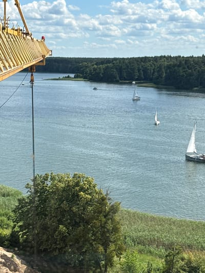 This media file depicts a wide shot of a body of water, likely a lake or bay, with a dense forest lining one side. Several sailboats are visible on the water, suggesting recreational sailing. In the foreground, a yellow crane structure extends from the left side of the frame, with a red light visible on its top. A body of water and a tree with green foliage are also visible to the right. The sky is partly cloudy. The overall scene suggests a pleasant, possibly sunny day. The image appears to be taken from an elevated perspective, possibly from the crane itself or a nearby elevated structure. No visible text is present in the image. The location context of Mikołajki, Poland, known for its lakes and sailing activities, aligns with the visual elements.