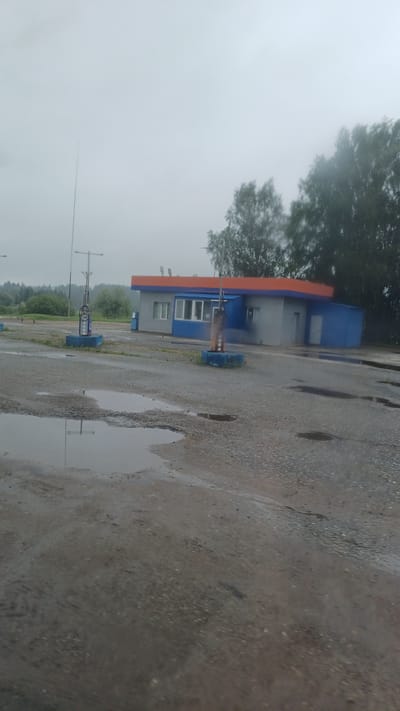 The image shows a small, abandoned gas station in the village of Kechevo, Russia. The building is painted blue and orange and appears to be in disrepair. There are two gas pumps in front of the building, both of which are also in disrepair. The ground in front of the gas station is covered in gravel and puddles, suggesting it has been raining. Trees and foliage are visible in the background. The sky is overcast and grey. There are no people or vehicles visible in the image.