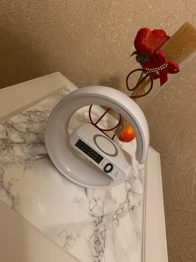 The image shows a white, crescent-shaped object with a digital display and a circular area on top. A small, red decorative flower with a red ribbon and gold beads is attached to the object by a red sparkly wire. The object is positioned on a surface with a marble-like pattern. In the background, a textured wall is visible. The scene is indoors. There is no information to indicate the location in the city of Kapan, Armenia.