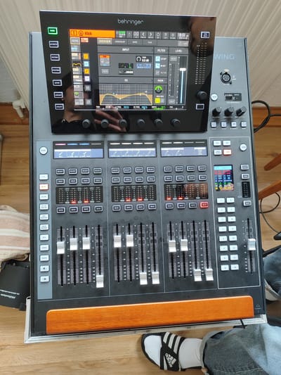 The image shows a Behringer Wing digital mixing console sitting on a table or surface indoors. The console's screen displays a channel labeled "Kick," with various settings visible, including EQ and input parameters. The physical interface features faders, buttons for channel selection, mute, and solo controls.

The lower right corner of the image shows a person's foot wearing an Adidas sandal with white socks. Next to the console on the left is a Millennium DI-33Aactive DI-Box.

While the image does not provide enough information to determine the location in the city of Göttingen, Germany.