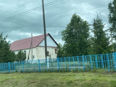 The image shows a building with a red roof and white brick walls, likely in Nadezhdino, Russia. A blue metal fence runs along the foreground, separating a grassy area from the building. Several trees, including evergreens and deciduous trees, are visible behind the fence and to the sides of the building. Power lines crisscross the overcast sky above. The scene appears to be a rural or semi-rural setting.
