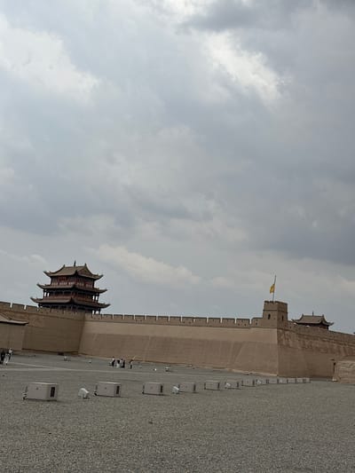 The image depicts a vast, fortified structure under a cloudy sky in Jiayuguan, China. The main subject is the Jiayu Pass, the western terminus of the Great Wall of China. The pass features a prominent multi-tiered pagoda-like structure with red accents on its roof and a dark tiled exterior, situated atop a high, tan-colored wall. To the right of the pagoda, a square watchtower rises from the wall, flying a yellow flag. Further to the right, another, smaller pagoda-like structure is visible. The wall itself is extensive and appears to be constructed of earth and brick. In the foreground, a wide, gravel-covered courtyard stretches out, with several white, rectangular electrical boxes or lighting units lined up. A few individuals, dressed in casual attire, are scattered throughout the courtyard, some walking and others standing in small groups. The overall impression is one of historical grandeur and a potentially overcast day.
