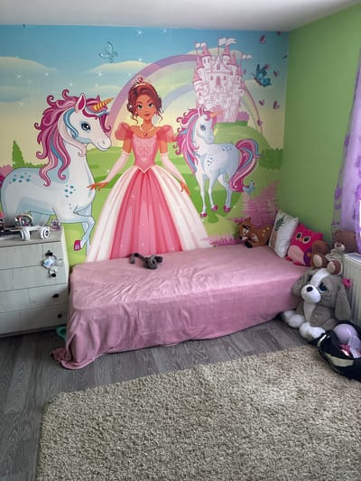 A child's bedroom is depicted. The walls are painted with a mural featuring a princess, unicorns, and a castle. A pink-covered bed is in the center of the frame with several stuffed animals arranged on the head. A rug lies on the floor. A white nightstand sits next to the bed. The location is Aldomirovtsi, Bulgaria. There are no people in the image.