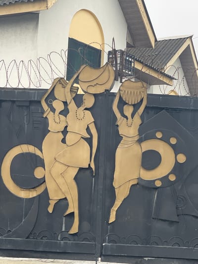 This image displays a dark gray or black metallic gate, likely an entrance to a property, adorned with prominent gold or dark tan-colored relief sculptures and abstract designs. The scene is set outdoors, with an overcast sky visible above. **People/Subjects:** The primary subjects are four stylized female figures, depicted in profile and relief, carved into the gate panels. Three figures are on the left panel, and one on the right. All figures wear short-sleeved tops and skirts or wrappers, and each has a visible beaded necklace. They are depicted performing the action of carrying various objects on their heads. The leftmost figure on the left panel carries a small, indistinct round object. The middle figure on the left panel carries a large, flattened, possibly woven basket. The rightmost figure on the left panel and the sole figure on the right panel each carry a large, round, fluted pot or basket on their heads, supported by raised arms. **Objects:** * **Gate:** A double-leaf metallic gate, predominantly dark gray/black with gold/tan relief work. The relief designs include the female figures, large abstract C-shaped and circular motifs, and smaller geometric patterns, including a series of semi-circular waves along the bottom edge. * **Security Wire:** Coiled razor wire, dark reddish-brown in color, is strung along the top of the gate and extends onto the walls of the background buildings. * **Building Elements (Background):** Sections of a building are visible behind the gate and wire. These include a white wall, a prominent arched recess or window frame in a light yellow/tan color, and a dark green-tinted window within. To the upper right, a section of a dark gray corrugated roof and light brown/tan soffits/eaves are visible, suggesting a pitched roof structure. **Actions/Interactions:** The stylized female figures are depicted in a static pose, illustrating the common practice of carrying goods on their heads. This action is culturally significant in many parts of Africa, including Nigeria. The figures and abstract designs are integrated into the gate, acting as decorative and potentially symbolic elements. The razor wire above indicates a security measure for the property. **Environment:** The image captures an urban environment, specifically identifiable as Lagos, Nigeria, given the architectural styles (such as the pitched roofs and building finishes), the prevalent use of decorative metallic gates with culturally relevant motifs, and the common application of razor wire for property security. The lighting suggests an overcast or muted daylight