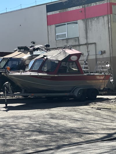 Here is a description of the image:

The image shows a red and gray motorboat on a trailer parked next to a building in Maple Ridge, Canada. The boat has the words "Thunderjet" and "Steelhead" visible, and a license plate number of "BC5342051" on the side. The building has a red stripe near the top and a couple of windows. The location is near the intersection of 224th Street and Lougheed Highway, Maple Ridge, Canada, based on the license plate number, the type of boat, and the surrounding environment. The ground is covered with asphalt. The photo appears to be taken outdoors in daylight.