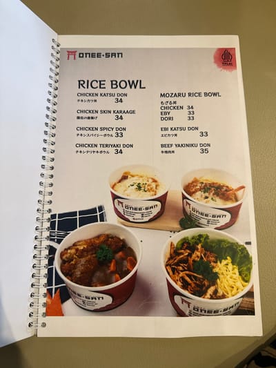 The image captures a close-up view of an open page from a spiral-bound menu, likely in a restaurant located in Kecamatan Cisaat, Indonesia. The page features various "RICE BOWL" and "MOZARU RICE BOWL" options from the brand "ONEE-SAN," whose logo is visible. A "HALAL" certification mark is displayed at the top left, indicating compliance with Islamic dietary laws.
The menu presents several dishes with both English and Japanese text, along with their prices. Featured items include "CHICKEN KATSU DON" (34), "CHICKEN SKIN KARAAGE" (34), "CHICKEN SPICY DON" (33), "CHICKEN TERIYAKI DON" (34), "CHICKEN" (34), "EBY" (33), "DORI" (33), "EBI KATSU DON" (33), and "BEEF YAKINIKU DON" (35). Each food item is accompanied by an appetizing photograph of the dish served in a distinctive white and red paper bowl. The images show a variety of toppings, such as fried chicken cutlets, crispy chicken skin, curry, stir-fried beef, and fresh greens.
The activity depicted is the presentation of food choices to a customer. The menu lies flat on a neutral-colored, possibly light brown or beige, surface, which could be a table or counter. The overall impression is that of a casual eatery offering a range of affordable and halal-certified Japanese rice bowls.