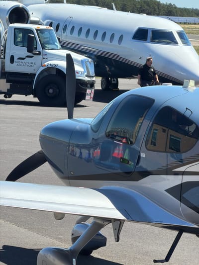 A white Signature Aviation truck is parked next to a small, grey propeller plane. The truck has the number "71257" printed on its fender. The small plane has a black propeller and a grey fuselage with black and white accents. In the background, a large white jet plane is visible. A man in a black shirt is walking away from the jet plane. The scene appears to be an airport tarmac, possibly in the Town of Southampton, United States of America, given the presence of aircraft and ground support vehicles. The sky is clear and blue, and the sun is casting shadows on the tarmac.