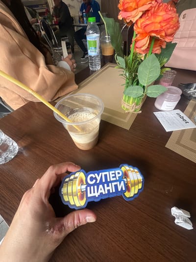 The image shows a table setting in what appears to be a cafe or restaurant in Batoshevo, Bulgaria, featuring a variety of objects, including artificial flowers, a beverage in a clear plastic cup with a yellow straw, a bottle of water, a Vanilla-scented candle, a betting slip from "inbet", and a sticker with Cyrillic text and an image resembling a dumbbell. A person's hand is holding the sticker, suggesting an interaction with the objects on the table. The setting appears to be indoors, with elements of daily life and leisure activities present.