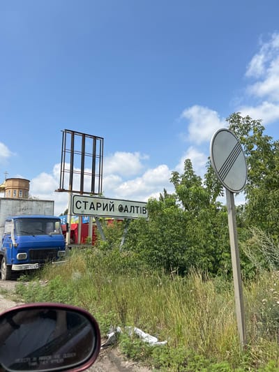The image depicts an outdoor scene located in Staryi Saltiv, Ukraine, under a bright blue sky with scattered white cumulus clouds.
In the left background, the upper part of a light-colored building with a golden onion-domed roof, indicative of a religious structure, is visible. To its right, a blue Soviet-era truck, likely a GAZ model, is parked or stationary. The truck's front bumper displays a partial license plate, "H 264-32XA." Above and slightly behind the truck, a tall, empty metal billboard frame stands.
Centered in the mid-ground, a white rectangular road sign with black Cyrillic lettering clearly reads "СТАРИЙ САЛТІВ." To the right of this sign, dense green foliage, comprising trees and bushes, occupies the middle and right sections of the frame. On the far right, a pole supports a circular white road sign with four parallel black diagonal lines, signifying the end of all local restrictions.
The immediate foreground consists of a dirt and gravel surface with overgrown green grass and weeds. A discarded white object is partially visible on the ground near the bottom center. A maroon rearview mirror from the photographing vehicle occupies the lower left corner of the frame, reflecting a distorted image of an arm or torso, indicating the presence of a person inside the vehicle.