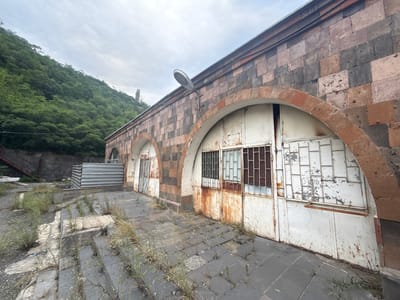 The image depicts an abandoned industrial building with arched entrances, located in the city of Kapan, Armenia. The building's exterior is constructed from rough-cut stone, with a mix of dark and reddish-brown hues. Two large, white metal doors, showing significant rust and peeling paint, are visible within arched openings. These doors have gridded windows. A smaller, similar door is visible to the left. The ground in front of the building is paved with dark stone tiles, cracked and overgrown with weeds and sparse grass. To the left, a set of concrete stairs leads upwards towards a dark stone wall and a metal structure. Beyond the building, a densely wooded hillside rises under an overcast sky. A single street lamp is mounted on the building's facade, near the roofline. The overall scene conveys a sense of neglect and disuse.