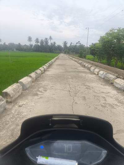 This image depicts a forward-facing view from a moving vehicle, likely a motorcycle, on a paved road in Barokulamgajah, Indonesia. The road is lined on both sides with a low, textured concrete barrier. To the left of the road, an expansive, lush green rice paddy stretches towards a cluster of palm trees and other foliage under a hazy, overcast sky. To the right, the road is bordered by a ditch and then by more trees and vegetation, with a utility pole and wires visible in the distance. The vehicle's dashboard is partially visible in the foreground, displaying a digital readout of "10" and a small blue indicator light. The overall scene conveys a sense of rural travel on a cloudy day.
