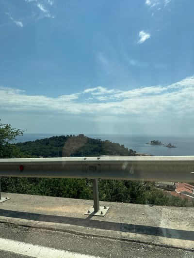 The image depicts a view from an elevated position in Brežine, Montenegro. A metal guardrail and concrete barrier dominate the foreground on the right side of the frame, bordering a road. Beyond the guardrail is a steep, densely forested slope leading down towards a body of water. In the distance, small rocky islands dot the sea. The sky is blue with scattered white clouds. A few houses with red tile roofs are visible amidst the trees on the right.