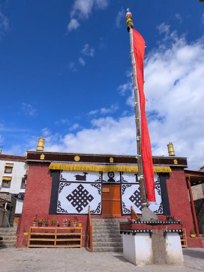 A bright red building with gold trim stands under a vibrant blue sky dotted with white clouds. A tall wooden pole with a long red prayer flag attached to it dominates the foreground. The building features decorative black and white panels on its facade, one with a symbol of a deer and another with an endless knot. There are wooden shelves filled with potted flowers placed in front of the building. Stone steps lead up to the building's entrance. The ground is paved with rough stone.