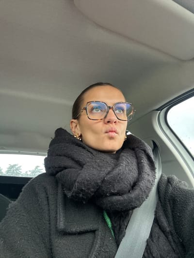 The image captures an adult female seated within a vehicle, presumably in the city of Saint-Cyprien, France. The subject is depicted from the chest up, with dark hair pulled back and wearing large, square-framed eyeglasses with tortoise-shell patterns and blue-tinted reflective lenses. Small, possibly gold-toned, clustered earrings are visible in both ears. Her lips are pursed into a 'pout' expression, and her eyes are directed upwards and slightly to the viewer's right. She wears a thick, black, textured scarf wrapped around her neck and a black, textured coat or jacket. A subtle green element is visible beneath the black coat near the collarbone. A grey seatbelt diagonally crosses her chest, extending from her right shoulder. The vehicle's light grey headliner is visible in the upper frame, and a blurred outdoor environment with an overcast sky and distant tree silhouettes is visible through the window on the left.