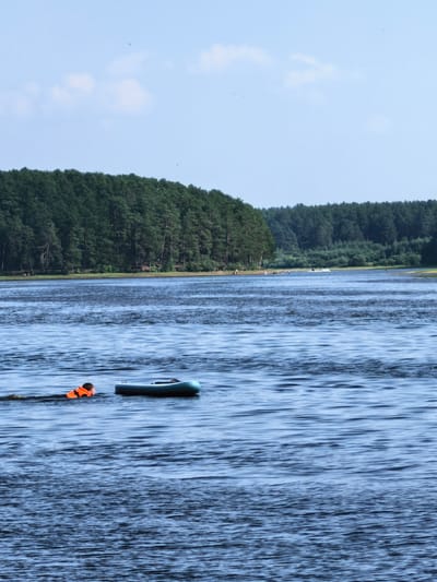 A person wearing an orange life vest swims in a dark blue body of water next to an inflatable boat. The person is on the left side of the frame and is swimming towards the right. The boat is also on the right side of the frame and is positioned horizontally. Behind the water, a dense green forest lines the shore, and beyond that, a clear blue sky with a few wispy clouds. The scene appears to be a recreational area, possibly a lake or river, located in the city of Sysert, Russia, given the presence of a forested area typical of the region and a body of water.