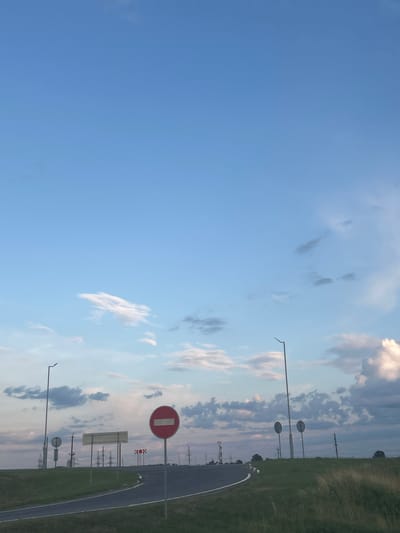 The image depicts a road with various traffic signs and infrastructure against a backdrop of a pale blue sky with scattered clouds. On the right side of the road, a prominent red circular sign with a white horizontal bar, indicating "No Entry," is visible. To the left, a series of smaller signs and a tall lamp post are situated on a grassy embankment. In the distance, power lines and pylons stretch across the landscape, suggesting a rural or semi-urban environment. The road curves to the left, leading into the distance. The scene appears to be captured during daylight hours, possibly late afternoon or early evening, given the soft lighting and cloud formations. The image does not contain any people or animals. The location is identified as Starosverzhansky Sel'skiy Sovet, Belarus.