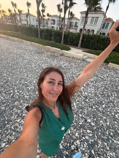 A woman is taking a selfie with her right arm extended. She is wearing a green sleeveless shirt and a silver necklace. The background shows a beach with pebbles, lush green bushes, and white houses with palm trees. The image appears to be taken during golden hour, with soft light illuminating the scene. The location is likely a residential area near a beach in Pervolia, Cyprus, given the architecture and vegetation.