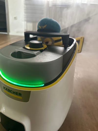 The image shows a close-up, eye-level view of a white and yellow Kärcher steam cleaner with a green light ring, placed on a wooden floor. A plush toy Minion wearing a teal beret with a golden emblem is perched on top of the steam cleaner, partially obscuring the control dial. Steam is visibly escaping from the device, creating a hazy effect in the background. The scene appears to be indoors, possibly in a domestic setting. There is no information available about the location in Heinebach, Germany.