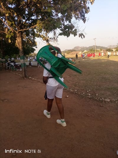 A person is depicted from behind, walking on a reddish-brown dirt path, carrying a bright green plastic chair upside down on their back. The individual wears a white t-shirt, white shorts, white sneakers, and white socks with green stripes. The text "KRPC FC" is visible on the backrest of the green chair.

The scene is an outdoor gathering, likely in the late afternoon given the clear sky and soft, bright light. On the left side of the image, a large, leafy tree partially shades a group of people dressed in white, seated on similar plastic chairs. To the right, beyond the dirt path, is a wide grassy field where a larger group of individuals, many in red uniforms, are gathered, possibly participating in an organized event, training, or formation. Several other figures are also present on the field. In the far background, distant hills and structures are visible under a pale blue sky. The general landscape suggests a rural or semi-urban environment in Ayere, Nigeria.

The visible text also includes an "Infinix NOTE 8i" watermark at the bottom left of the original image, indicating the camera used.