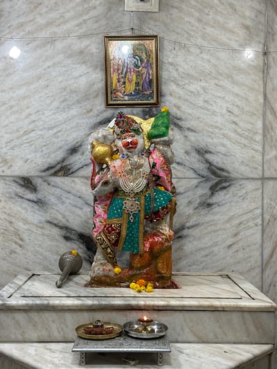 Captured indoors, the image displays a statue adorned in vibrant textiles and jewelry, set within what seems to be a temple or shrine. The backdrop is a marble wall exhibiting natural veining. To the statue's left hangs a framed picture. To its right, a religious metal ornament is mounted on the wall, next to an enclosed corner. A silver metal spouted jug lies nearby on the floor. This site is in Vadodara, India.
