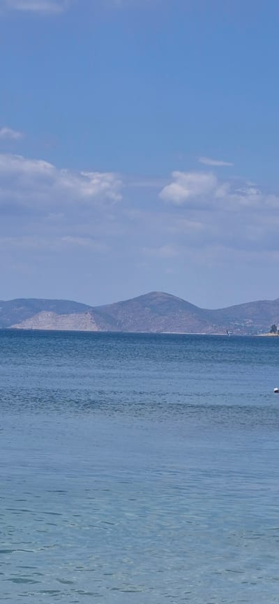 The image depicts a serene seascape with a clear blue sky dotted with white clouds. In the distance, a range of barren, brown hills forms the horizon. The sea in the foreground is calm and transparent, revealing the sandy seabed beneath. There is a small sailboat visible on the water. A person wearing a white cap is partially visible in the water on the right side of the frame, seemingly engaged in swimming or water activity. The scene captures a peaceful moment at a beach, likely in Paleo Faliro, Greece, given the characteristic landscape of the Attica coastline.