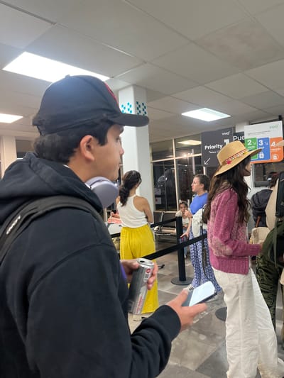 Here is a description of the image:
The image shows a slightly rotated, close-up view of a crowded airport terminal. In the foreground, a person wearing a black baseball cap, black hoodie, and gray headphones is holding a can of soda and a smartphone, seemingly taking a picture. The foreground subject is predominantly out of focus.
Further into the frame, several people are standing in a line, presumably waiting. One person wears a pink sweater, white pants, and a straw hat. Behind them, other people are wearing blue shirts and dresses.
Signs are visible in the background with the words "Priority" and "Viva," along with colored icons representing numbers 2, 3, and 4, with labels "verde," "azul," and "naranja" beneath them. Based on the signs in the image, the location is in the city of Puerto Escondido, Mexico.