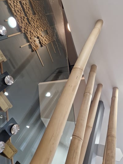 The image depicts an interior space with a modern aesthetic, featuring elements of natural materials and reflective surfaces. Prominently visible are several large, light-colored bamboo poles, angled upwards towards a white ceiling. Some of these poles appear to be structural supports, with visible nodes characteristic of bamboo.

On the left side of the image, a wall is covered with a textured, wood-like material arranged in a grid of small cubical blocks. A circular light fixture with a white diffuser is embedded in this wall. Adjacent to this wall is a large, highly reflective surface, likely glass, which mirrors parts of the bamboo poles and the ceiling, creating a sense of depth and spatial complexity.

Several circular, metallic fixtures with a brushed finish are attached to the reflective surface, appearing to hold or support other elements. Below these fixtures, wooden components that resemble shelves or display units are partially visible.

In the background, a dark grey angular structure is visible, possibly a pillar or part of the building's architecture. The overall lighting in the scene is soft, with some sources contributing to the reflections on the glass. The image does not contain any people.

This scene is located in the **Médiathèque La Canopée, Antony, France.**