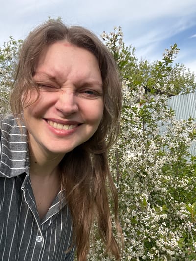 A selfie of a fair-skinned person with long brown hair, smiling and winking. The person is wearing a gray and white striped button-down shirt. Behind the person, there is a flowering tree or bush with white blossoms and green leaves, and a glimpse of a corrugated metal building in the background. The sky is visible in the upper left corner, with wisps of clouds. The specific location in Aksay, Kazakhstan, cannot be determined from the image alone.
