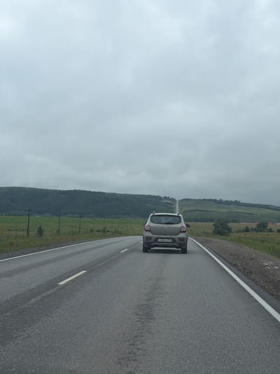 The image depicts a car driving on a wet asphalt road under an overcast sky. The car, a light-colored hatchback, is in the foreground, facing away from the camera and towards the left side of the frame. It has a license plate that reads "C356YH 102". The road has a white line marking the edge.
To the left of the road, a green field stretches out, bordered by a dense forest that climbs a hillside. In the distance, the landscape appears hilly and covered with vegetation. The weather is gloomy and cloudy, suggesting it might be raining or has recently rained, as indicated by the wet road surface and the overall muted lighting.
The scene is set in a rural or semi-rural area, with the natural landscape dominating the background. The image appears to be taken during daylight, though the heavy cloud cover obscures any direct sunlight and contributes to the somber mood. There are no people visible in the frame. The overall impression is one of a solitary journey through a quiet, natural landscape on a cloudy day. The location context provided is Анновка, Russia.