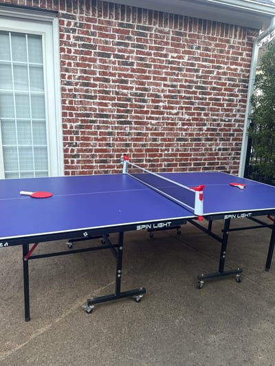 The image depicts a Spin Light brand ping pong table set up on a concrete surface, likely a patio or driveway. The table is located next to a brick building with a white-framed window. The ping pong table has a net and two paddles on its surface. The scene is located in Southlake, United States of America.