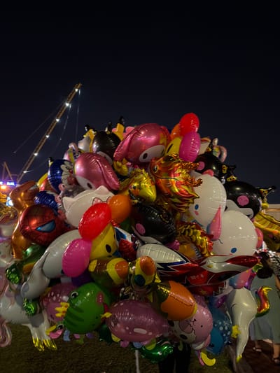The image depicts a dense cluster of numerous inflated Mylar balloons, predominantly featuring various cartoon characters, animals, and generic shapes, displayed or carried outdoors at night in An Hải Ward, Vietnam. The balloons include specific characters such as Spiderman, Hello Kitty, My Melody, Mickey Mouse, a black-and-white cow, and a diverse array of fish, frogs, unicorns, elephants, and other animals. They exhibit a wide spectrum of metallic and solid colors including pink, gold, red, blue, green, yellow, orange, black, and white.

A person, partially visible at the bottom center in dark attire, appears to be holding the large collection of balloons. Other indistinct human figures are present in the dimly lit lower right background. The immediate ground beneath the balloons is dark and textured. The background reveals a dark, clear night sky. In the upper left quadrant, a tall construction crane, illuminated by multiple white lights along its structure, is visible against the dark sky. Artificial light sources from an urban or commercial setting illuminate the scene, casting reflections on the metallic balloons.