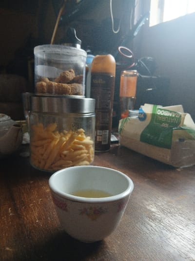A table displays assorted items: two glass jars, one containing yellow snack sticks and another holding brown sticks. Several bottles are positioned above them, with a partially visible, brown bag in the background. A patterned teacup is also visible to the right of the snack jar, on the table surface.