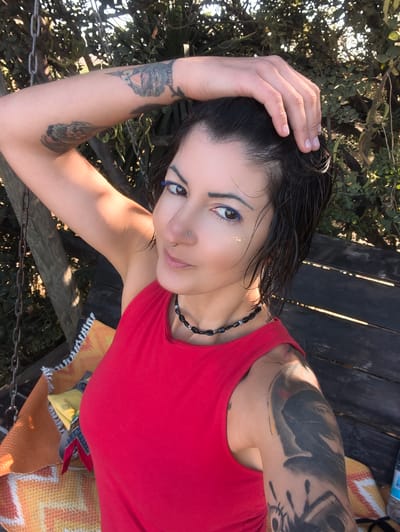 The image captures a female individual positioned outdoors in the city of Shaalvim, Israel. She is centered in the frame, looking directly at the camera with a slight smile. Her dark hair appears wet, and her right hand is placed on top of her head, with fingers extended. She wears a red sleeveless top and a black beaded necklace with interspersed blue beads. Tattoos are visible on both arms; the right arm displays a design resembling an owl or bird, while the left arm features a large, dark, abstract design. Her facial makeup includes blue eyeliner along the lower lash line and small, reflective, glitter-like particles on her right upper cheek. She has a silver stud piercing on her right nostril and a silver ring piercing on her septum. The background consists of dense green foliage, indicating an outdoor, possibly wooded, environment. Sunlight filters through the leaves, creating areas of brightness and shadow. To the left, a wooden structure and metal chains are visible, suggesting a swing or similar apparatus. To the right, a dark wooden slatted surface, likely a bench or seat, is partially covered by a woven blanket in shades of orange and yellow with white fringed edges. A small yellow object and a portion of a plastic bottle are also present on the wooden surface.